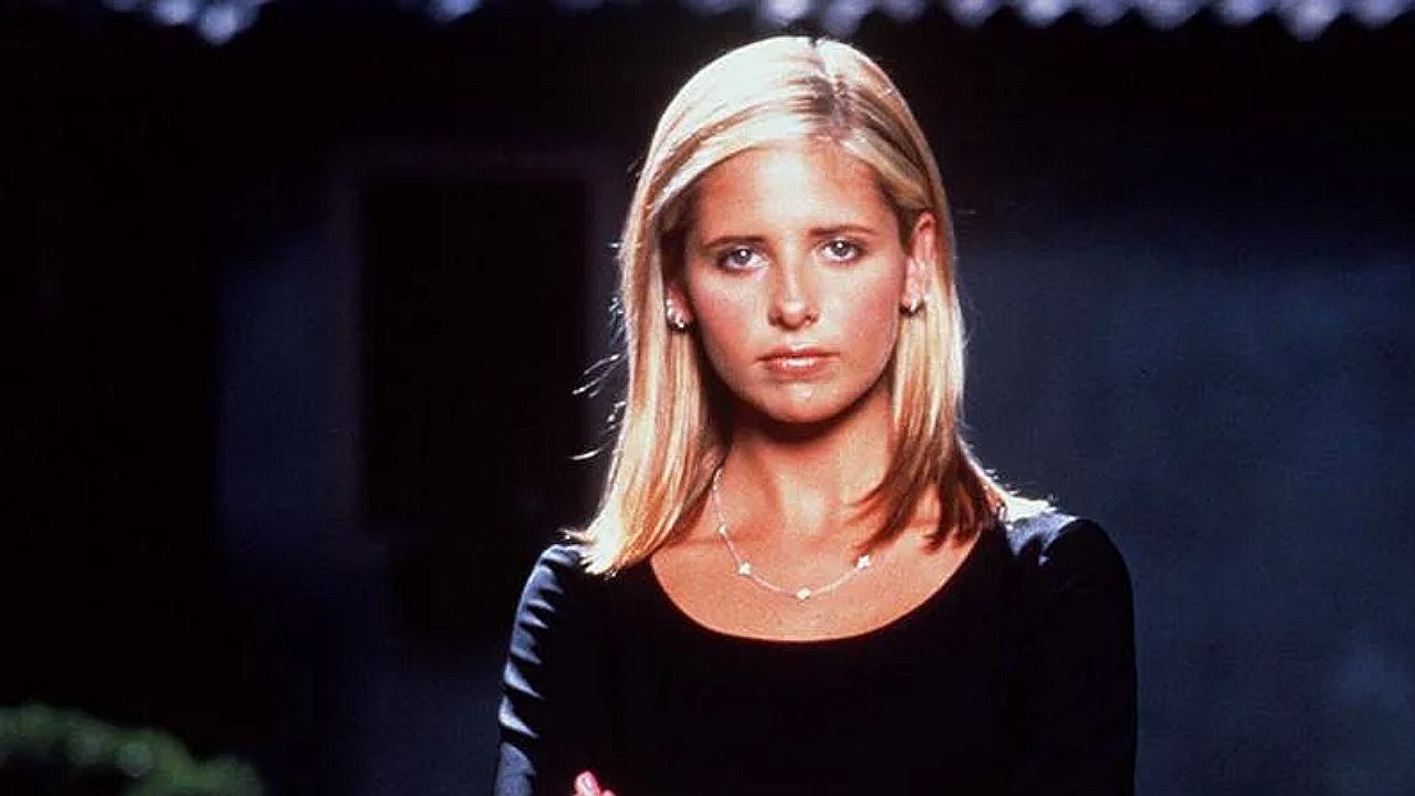 Uma mulher jovem de cabelo loiro, liso e na altura dos ombros está olhando diretamente para a câmera com expressão séria. Ela usa uma blusa preta de mangas compridas e um colar delicado. A iluminação destaca o rosto enquanto o fundo aparece escuro e desfocado, sugerindo uma cena externa à noite ou em ambiente pouco iluminado.