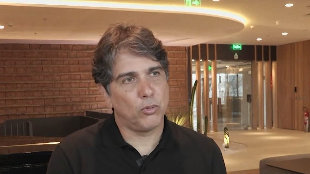 Um homem aparece em um ambiente interno moderno, durante uma entrevista. Ele veste uma camisa escura e tem cabelo curto com fios grisalhos. Sua expressão é séria e concentrada, olhando levemente para o lado enquanto fala. Ao fundo, há um espaço bem iluminado com paredes de tijolos aparentes, iluminação embutida no teto e uma porta de vidro, sugerindo um ambiente corporativo ou hotel.