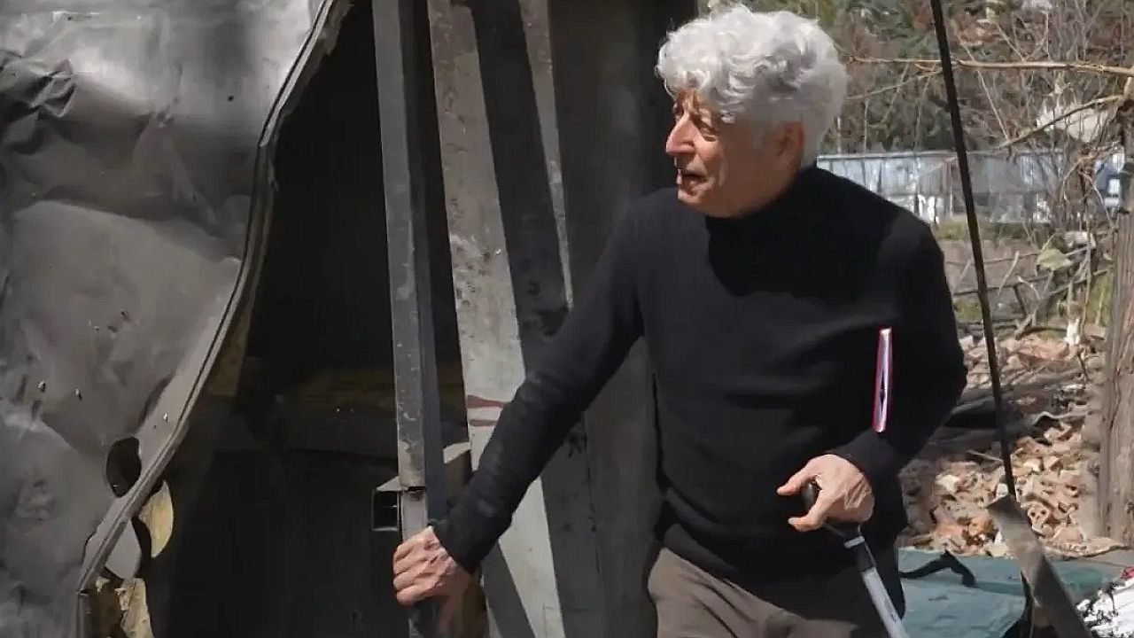 Homem idoso de cabelo branco segura bengala ao lado de barraco em área externa com entulho