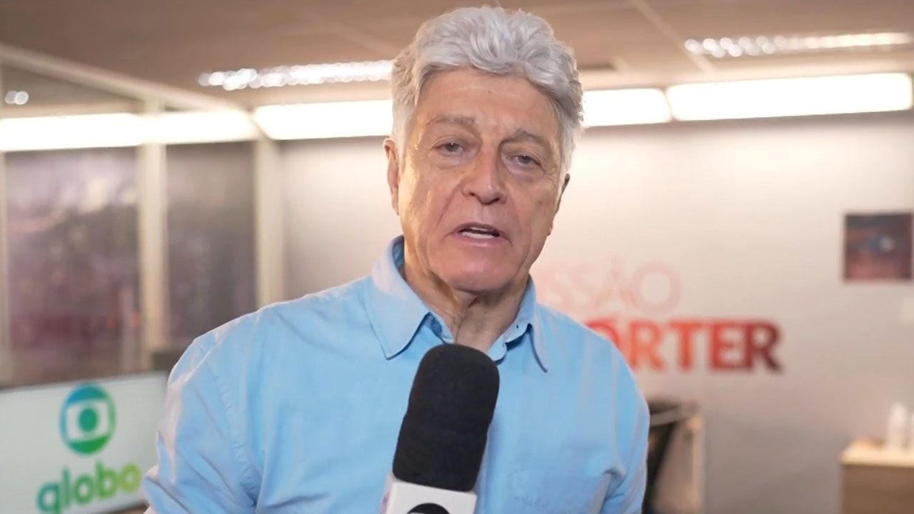 O jornalista Caco Barcellos de camisa azul clara fala ao microfone na redação do Profissão Repórter, com o logo da Globo ao fundo