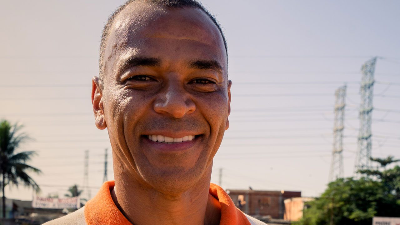 Ex-jogador Cafu sorri de camisa polo laranja em ambiente externo com torres de alta tensão e vegetação sob céu claro ao fundo