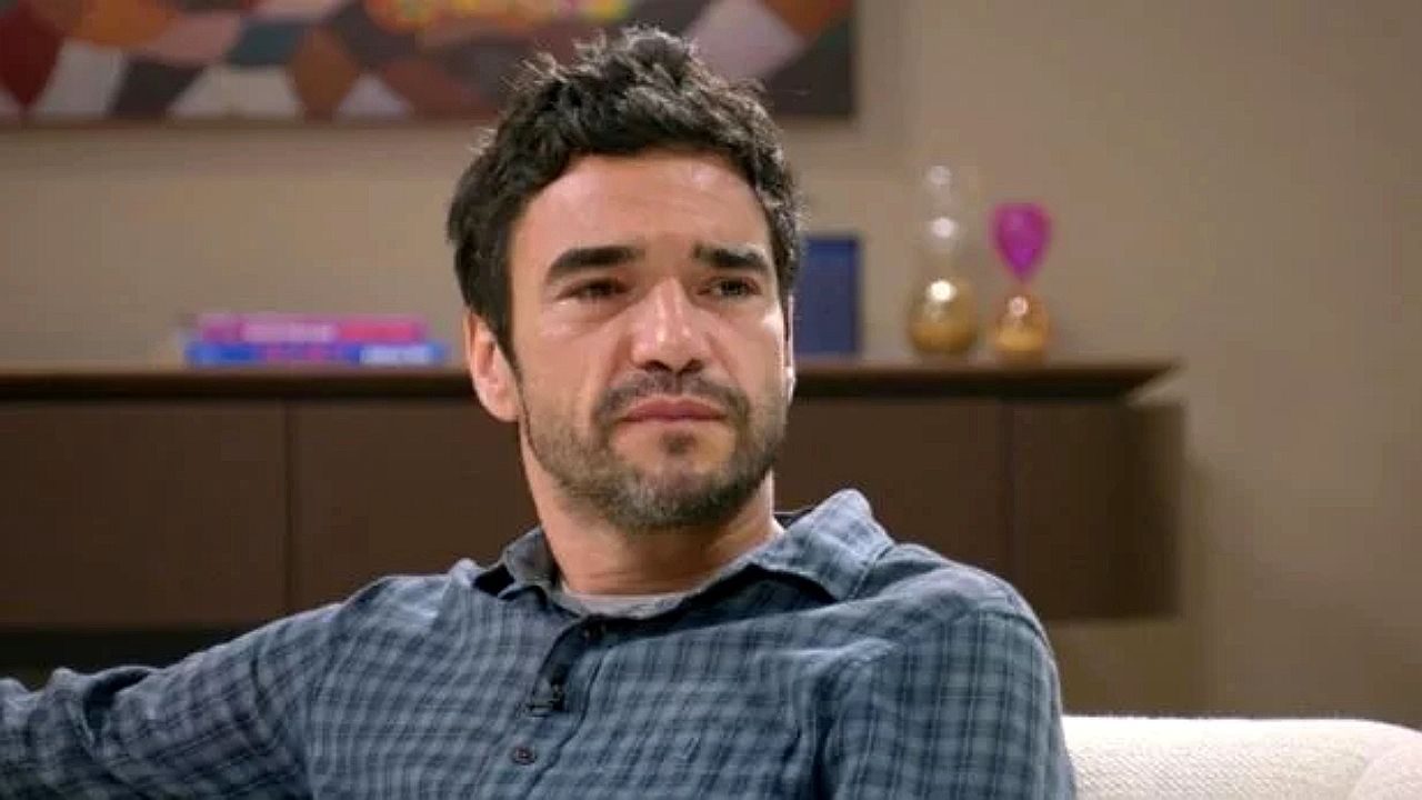 Homem de barba curta e camisa xadrez observa alguém com expressão séria e emocionada em sala de estar com móveis e objetos decorativos ao fundo