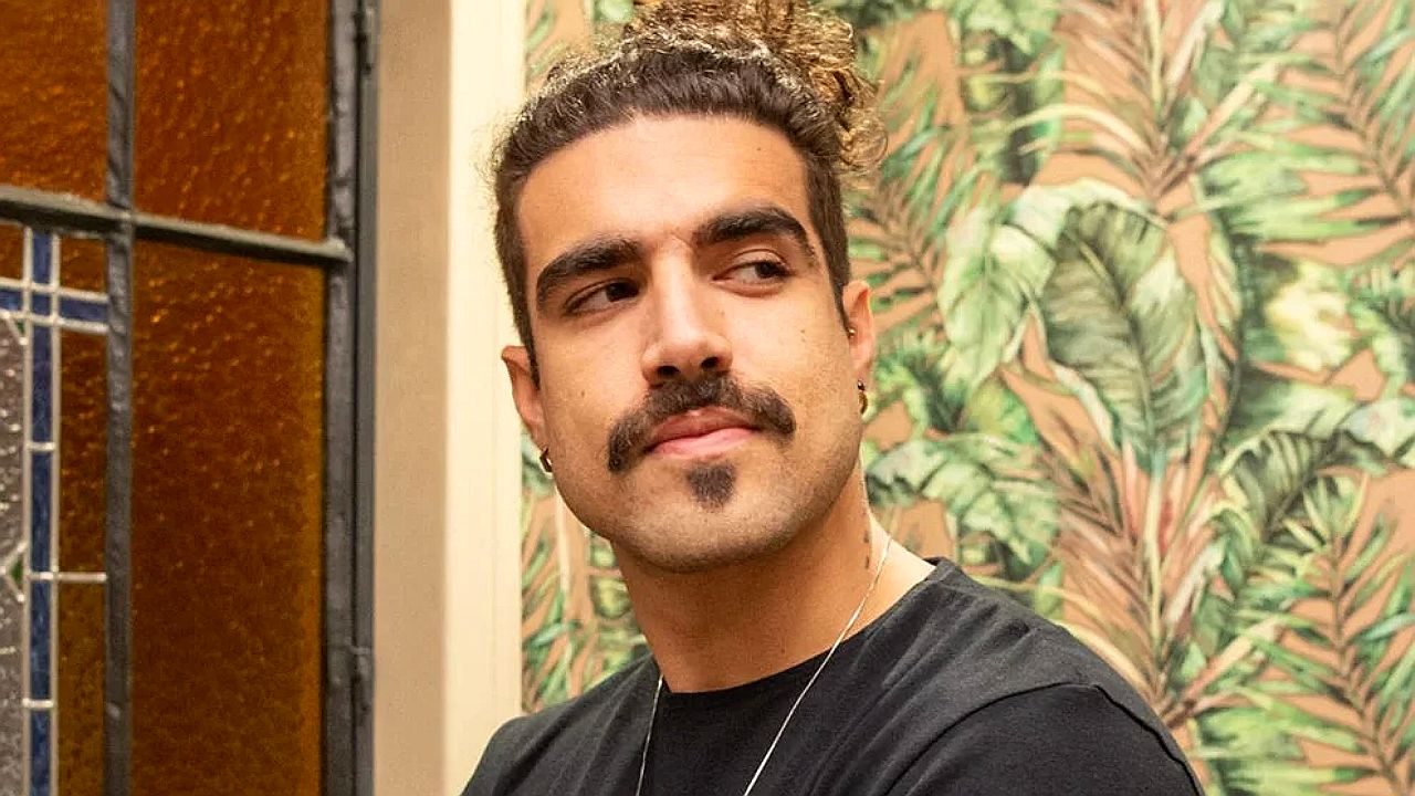 Homem de bigode e cabelo preso olha de lado diante de parede com estampa de folhas em ambiente interno