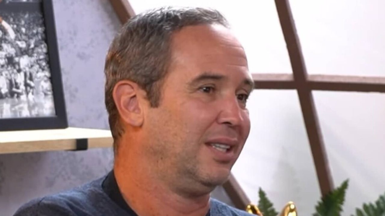 Homem de cabelo curto e barba rala fala em ambiente interno com prateleira e plantas ao fundo