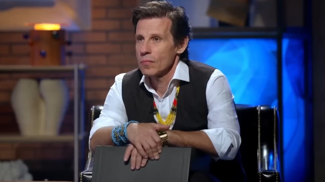 Caito Maia aparece atento e concentrado durante sua participação no Shark Tank Brasil. Sentado em uma poltrona no estúdio do programa, o empresário usa camisa branca, colete preto e acessórios marcantes, como pulseiras e colares coloridos de inspiração indígena. A iluminação azul ao fundo compõe o cenário característico do reality de negócios.