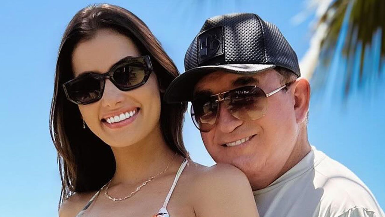 Calita Franciele sorri ao lado de Amado Batista que usa boné preto e óculos escuros em dia ensolarado ao ar livre