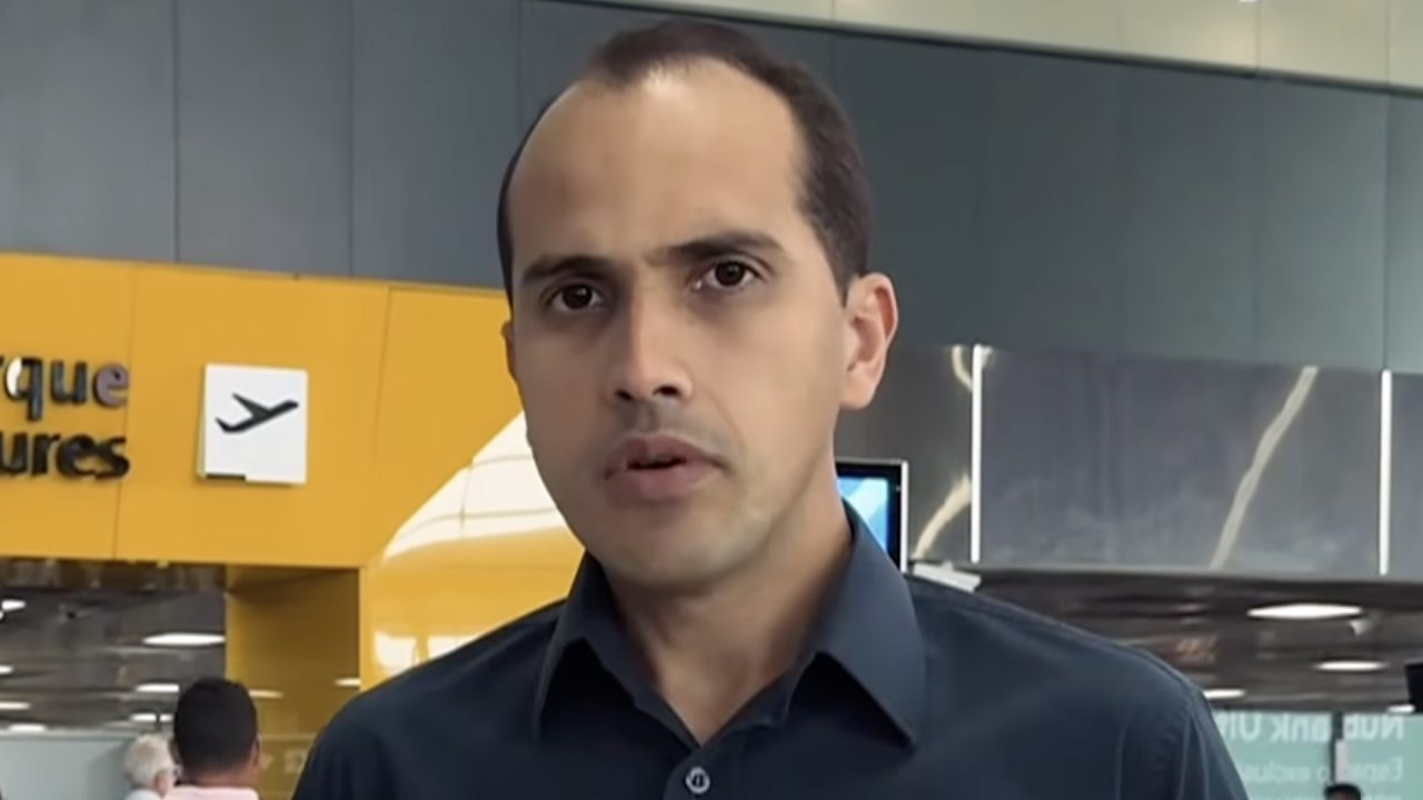 Repórter de camisa azul-escura fala à câmera no aeroporto de Guarulhos durante gravação do Câmera Record