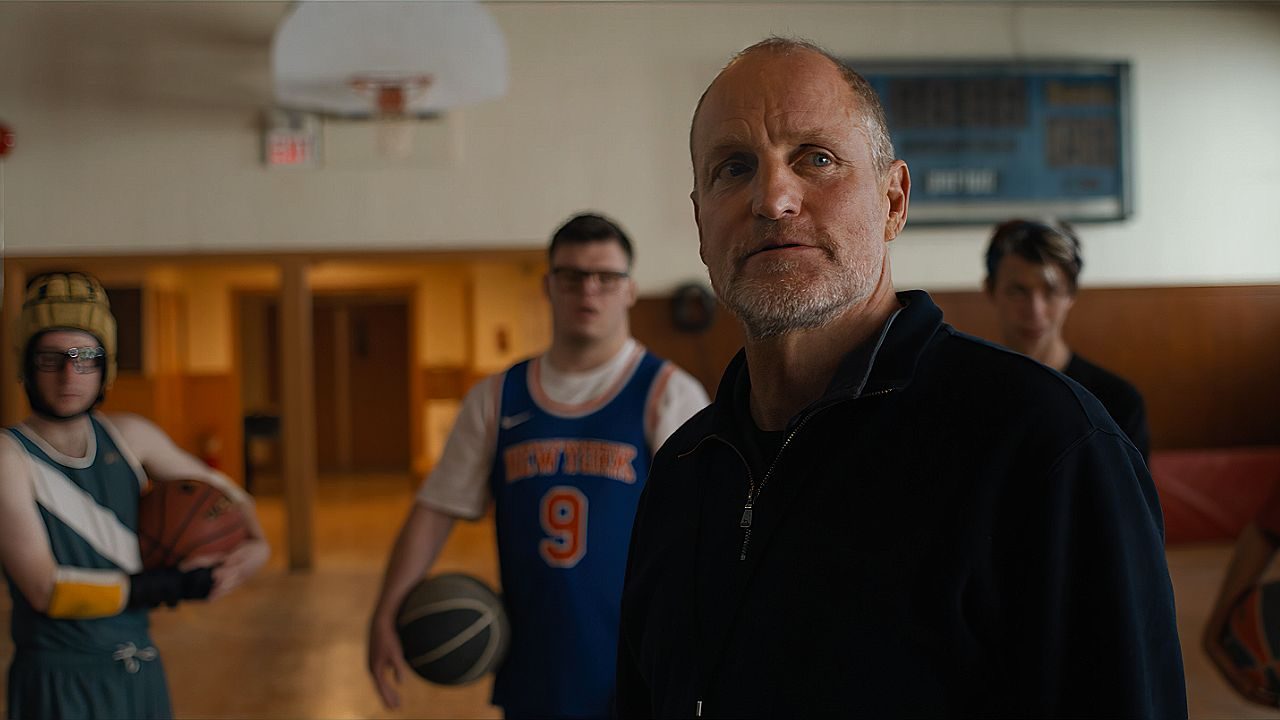 Homem mais velho, com expressão séria, está em uma quadra de basquete escolar, enquanto jovens ao fundo seguram bolas e vestem uniformes esportivos. O cenário sugere treino ou reunião tensa em ambiente esportivo interno.