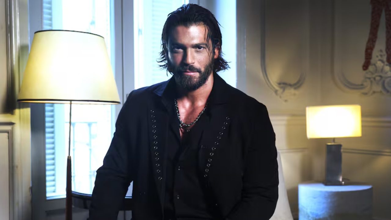 Homem com cabelo comprido penteado para trás e barba aparece sentado em um ambiente elegante e iluminado por luz natural e abajures. Ele veste camisa e blazer pretos com detalhes sutis e colares no pescoço, olhando diretamente para a câmera com expressão confiante. Ao fundo, há uma janela e decoração clássica em tons claros.