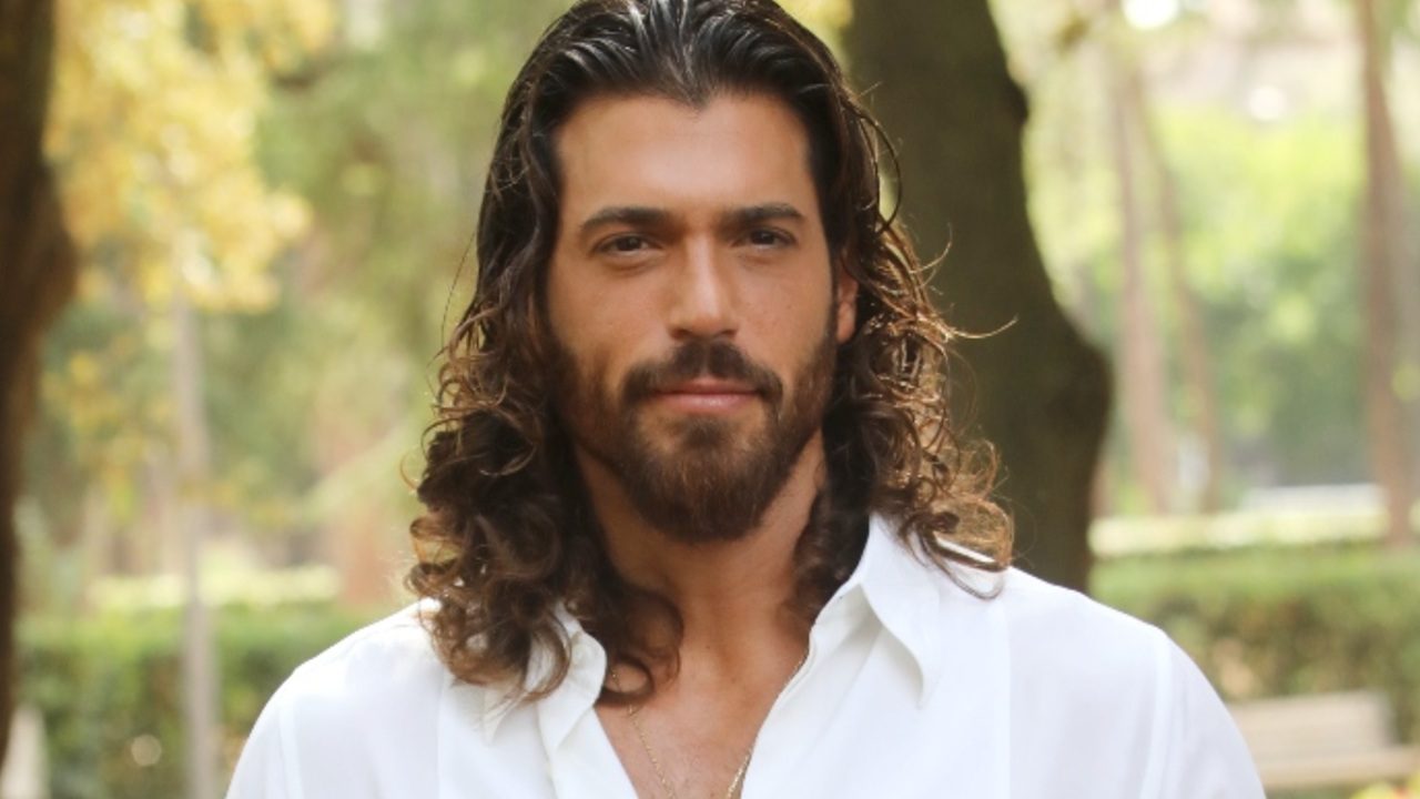 Homem de barba e cabelos longos e ondulados aparece em ambiente externo iluminado, vestindo camisa branca. Ele olha diretamente para a câmera com expressão serena; ao fundo, vê-se um parque com árvores e vegetação desfocada