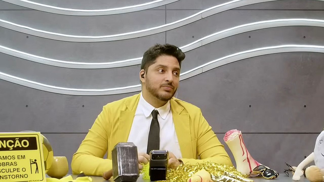 Homem de blazer amarelo sentado em bancada de programa com objetos variados e celular sobre a mesa