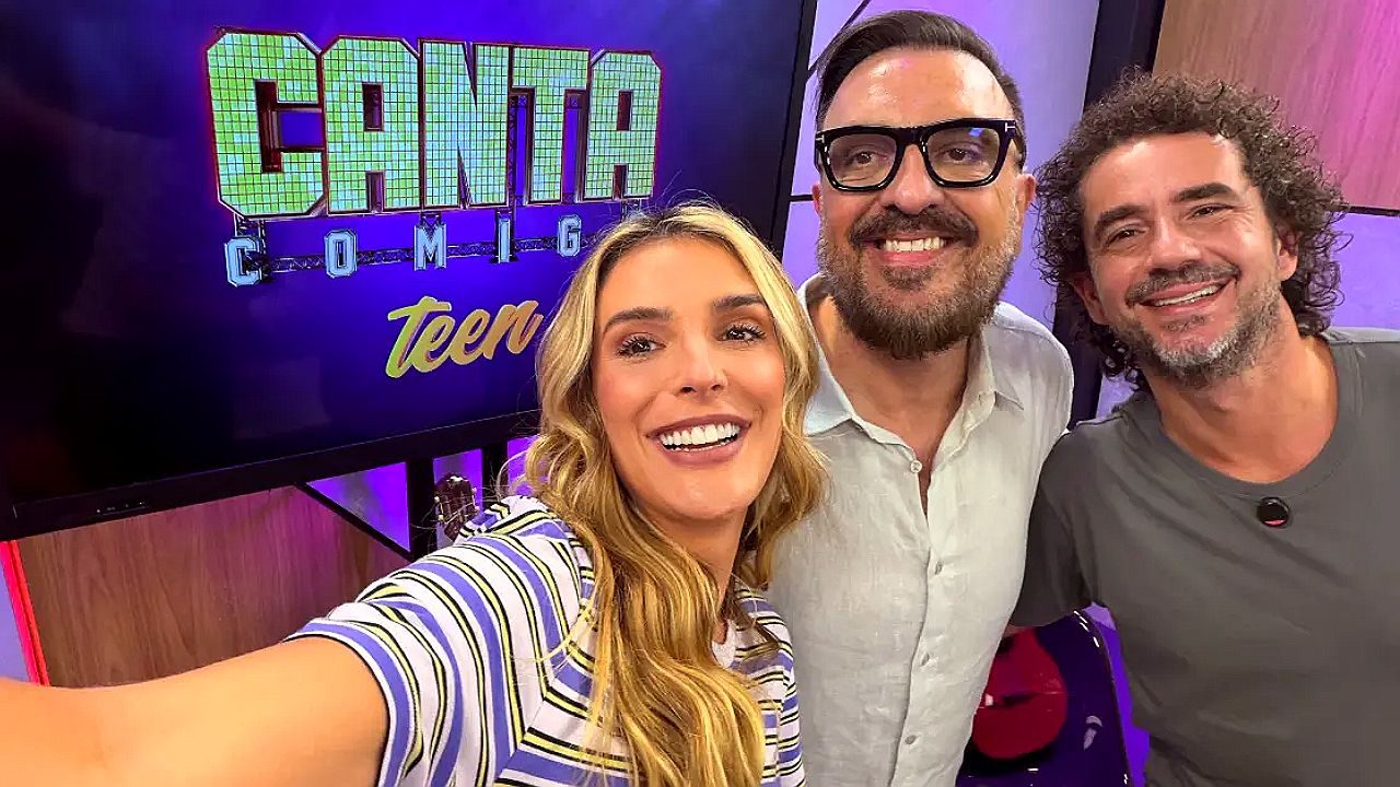 Apresentadores sorriem em selfie no estúdio com tela do programa Canta Comigo Teen ao fundo