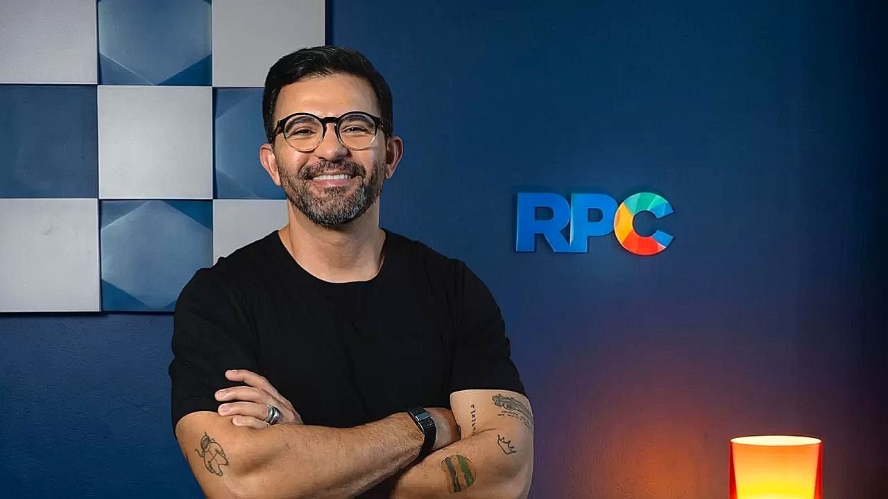 Apresentador sorri com braços cruzados em frente ao logotipo da RPC em estúdio da afiliada da TV Globo no Paraná