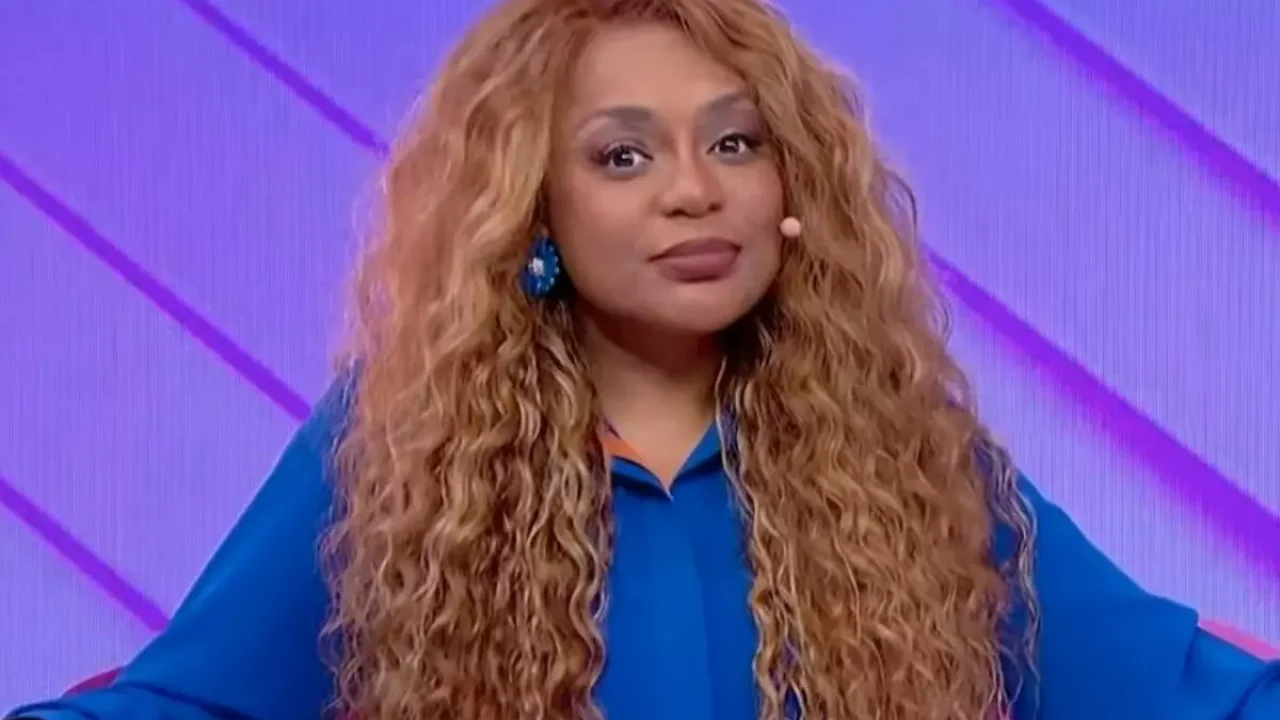 Cariúcha em programa de TV, sentada em poltrona rosa e usando camisa azul com microfone no rosto