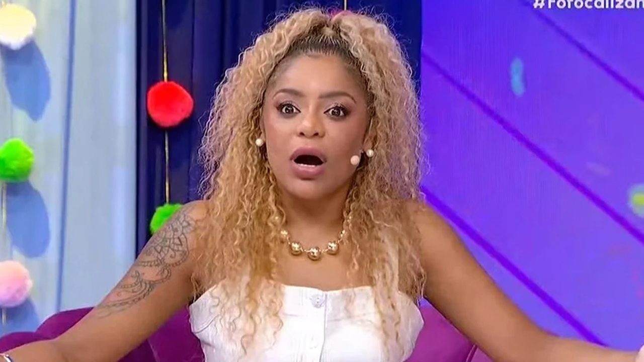 Mulher com expressão surpresa participa de um programa de TV colorido, vestindo roupa branca, colar dourado e cabelo cacheado loiro preso no alto.