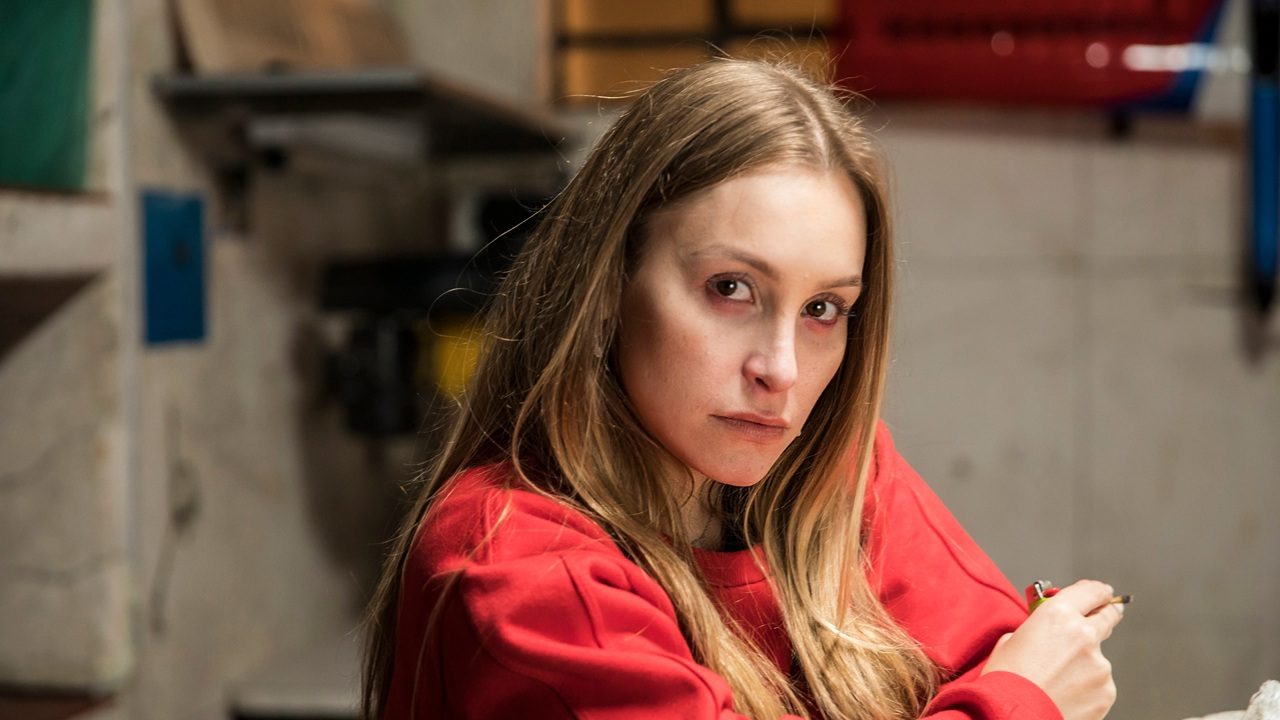 Mulher jovem de cabelos loiros e lisos, usando um moletom vermelho, olha diretamente para a câmera com expressão séria em um ambiente interno simples, com prateleiras e objetos desfocados ao fundo