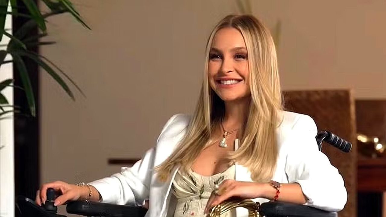 Carla Diaz sorri sentada em cadeira de rodas para cena de novela