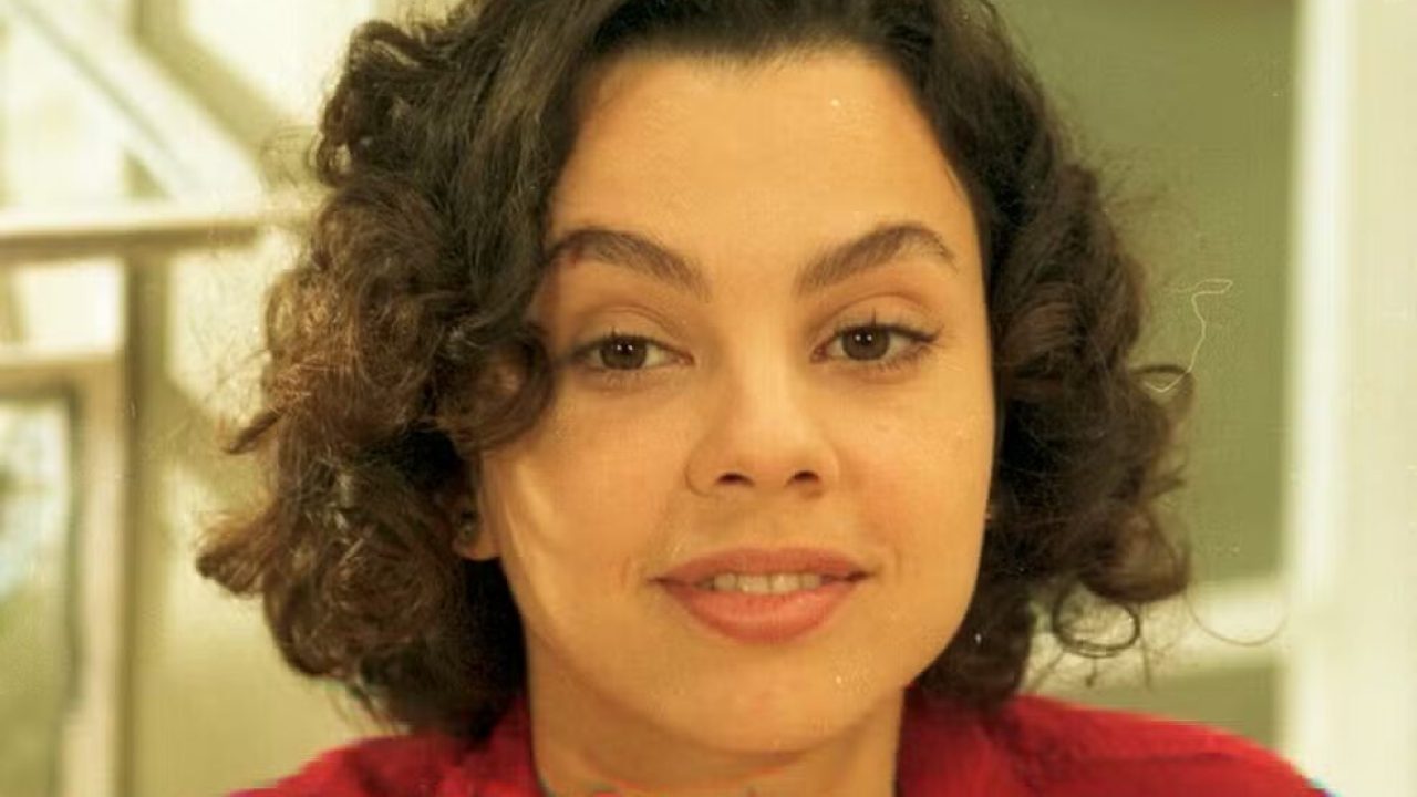 Carla Marins como Joyce em História de Amor, com expressão serena e figurino vermelho