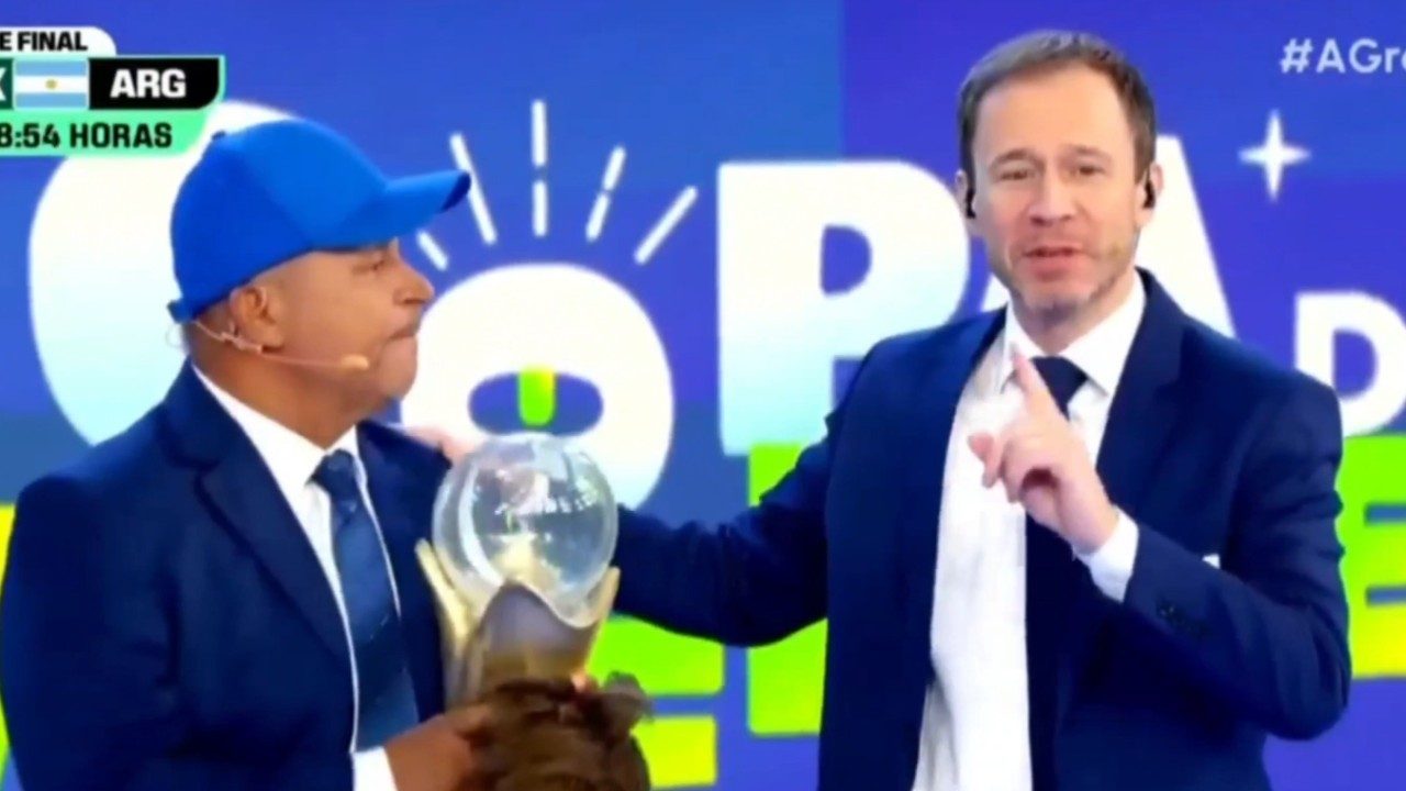 Carlinhos Vidente de boné azul segura troféu ao lado de Tiago Leifert de terno durante transmissão em estúdio