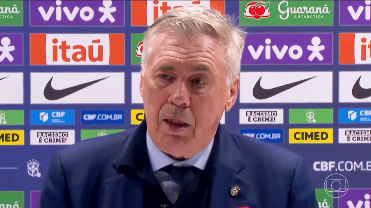 A imagem mostra Carlo Ancelotti, um homem de cabelos grisalhos durante uma coletiva de imprensa, em frente a um painel com logotipos de patrocinadores da Confederação Brasileira de Futebol (CBF), como Itaú, Vivo, Nike, Cimed e Guaraná Antarctica. Ele veste terno azul escuro com gravata e crachá da seleção brasileira, respondendo perguntas da Globo após a partida entre Brasil e Senegal