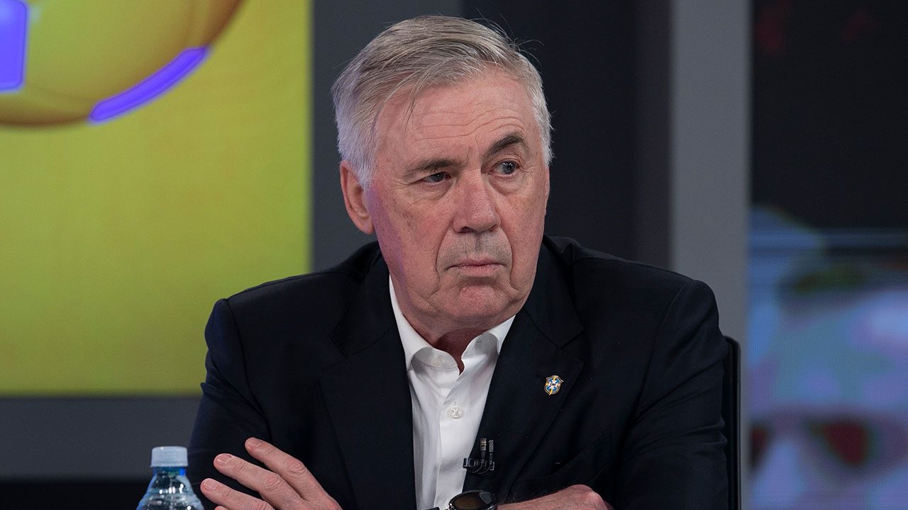 Técnico Carlo Ancelotti de camisa social branca e terno preto com olhar sério e braços cruzados durante entrevista para Record