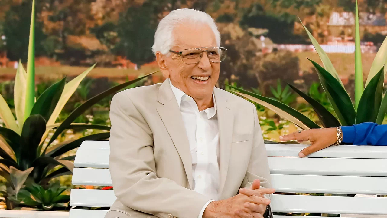 Carlos Alberto de Nóbrega sorrindo sentado em banco branco no cenário de A Praça é Nossa