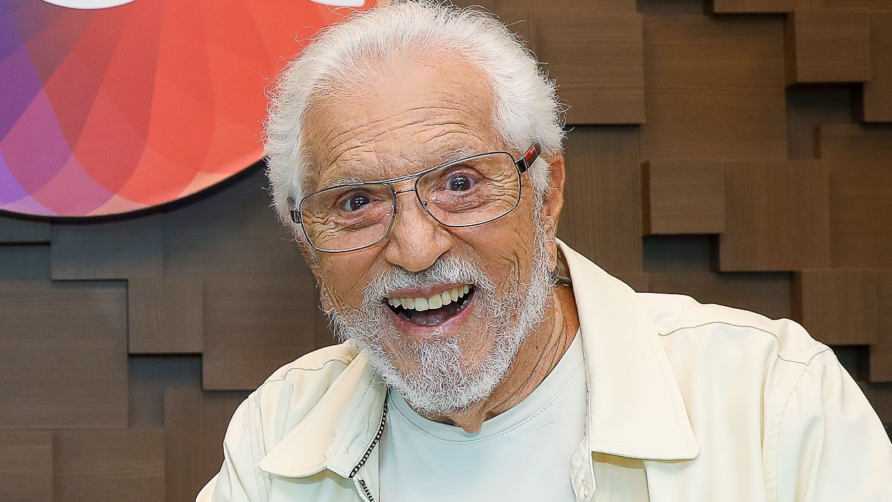 Carlos Alberto de Nóbrega sorri para a câmera, apresentador veterano de cabelos brancos e barba por fazer, veste jaqueta bege clara, fundo amadeirado