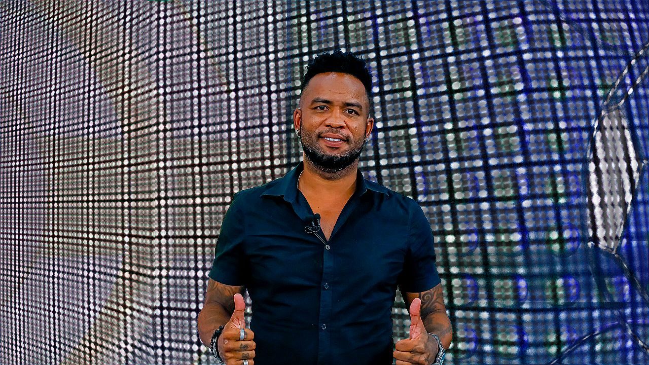 Homem em um estúdio de TV, usando camisa preta e microfone preso à roupa, sorrindo e fazendo sinal de positivo com os dois polegares.