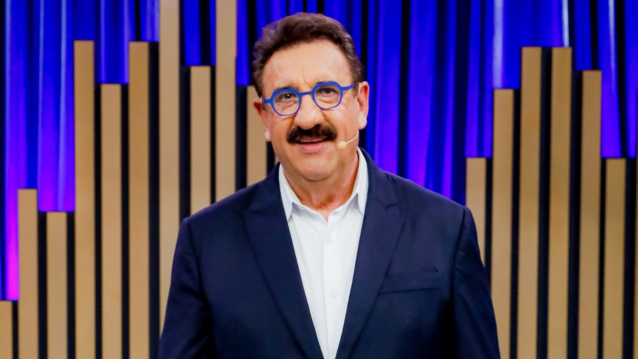 Ratinho apresenta programa em estúdio com fundo de painéis verticais iluminados usando terno escuro óculos azuis e microfone headset