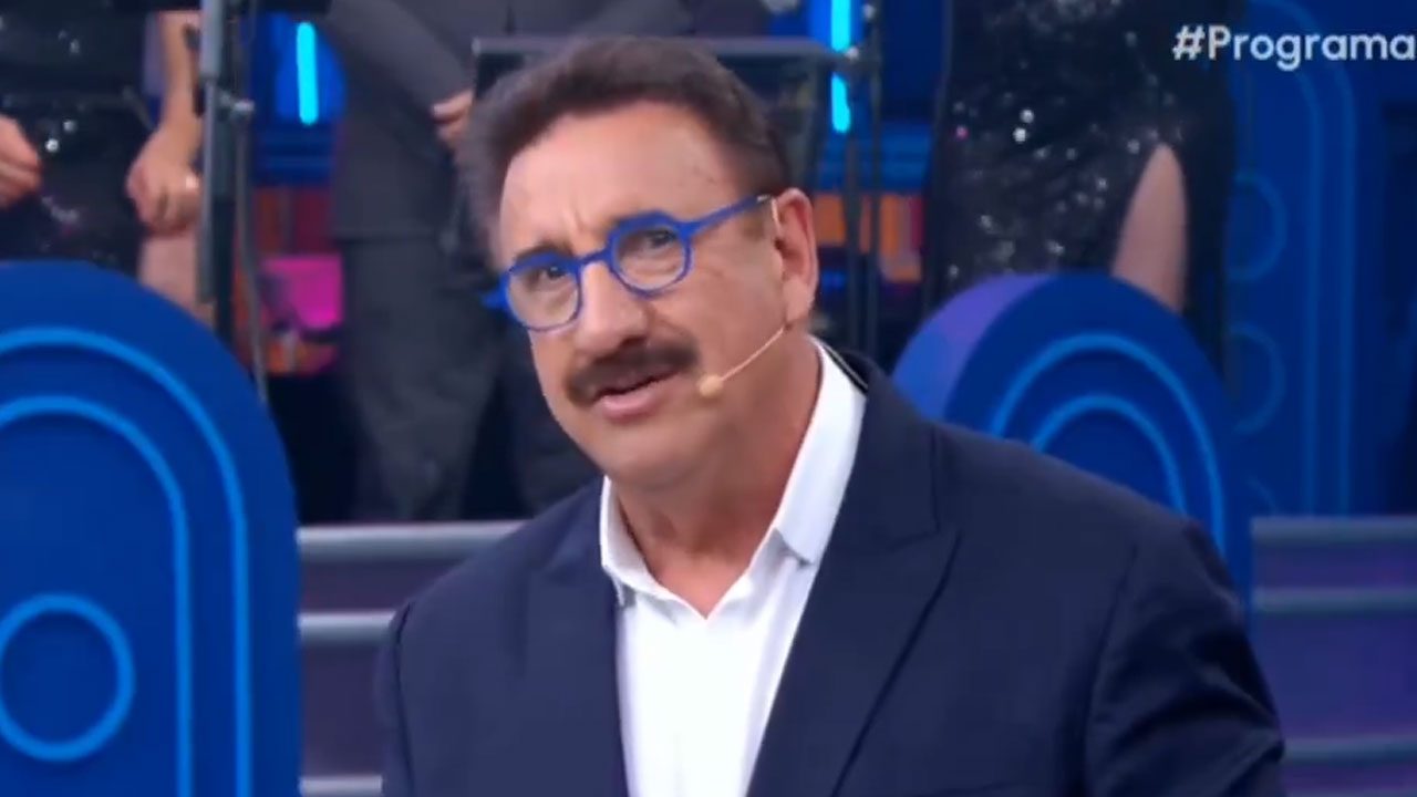 Apresentador Carlos Massa Ratinho com terno azul e óculos de aro azul no palco do seu programa no SBT com fundo roxo