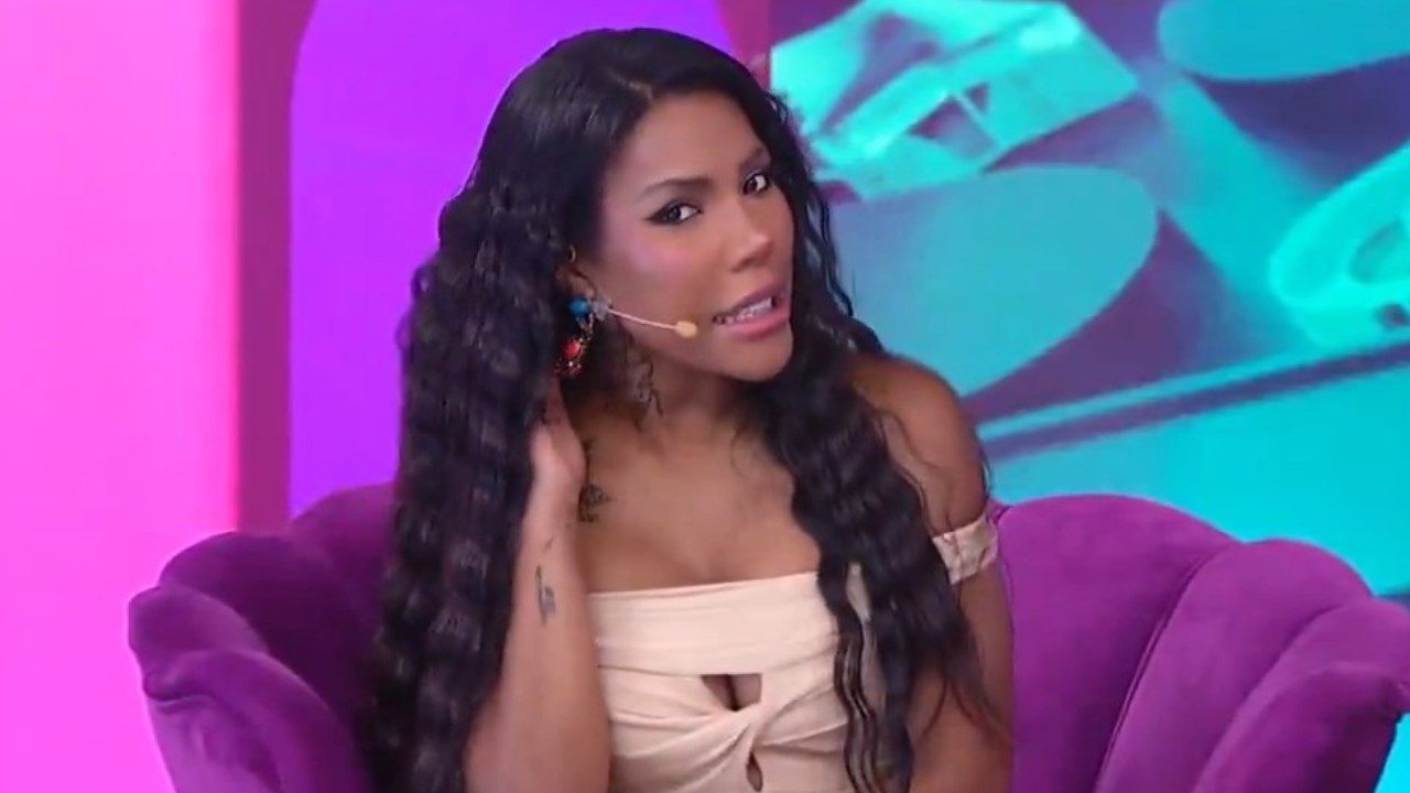 Carol Lekker no programa Fofocalizando do SBT, usa vestido bege decotado, sentada em poltrona roxa, longos cabelos escuros ondulados, cenário colorido ao fundo