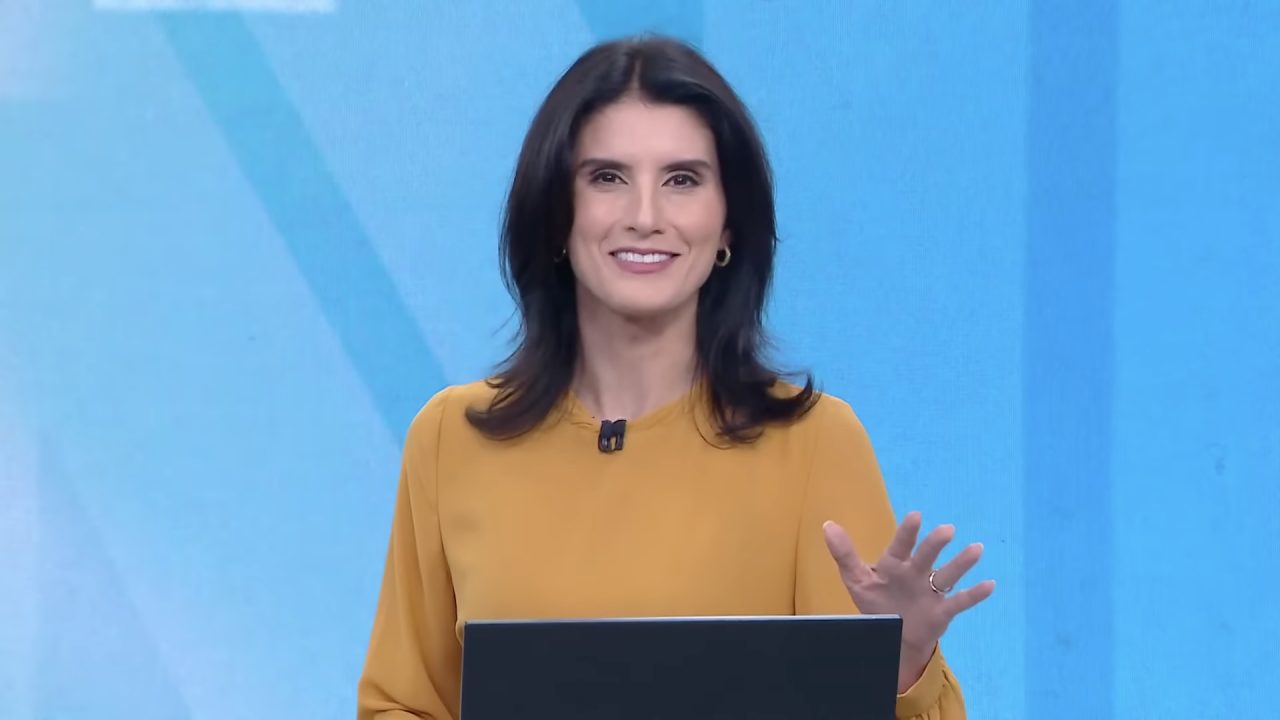 Carol Nogueira, de blusa amarela, sorri e gesticula diante de notebook no estúdio azul do Novo Dia na CNN Brasil