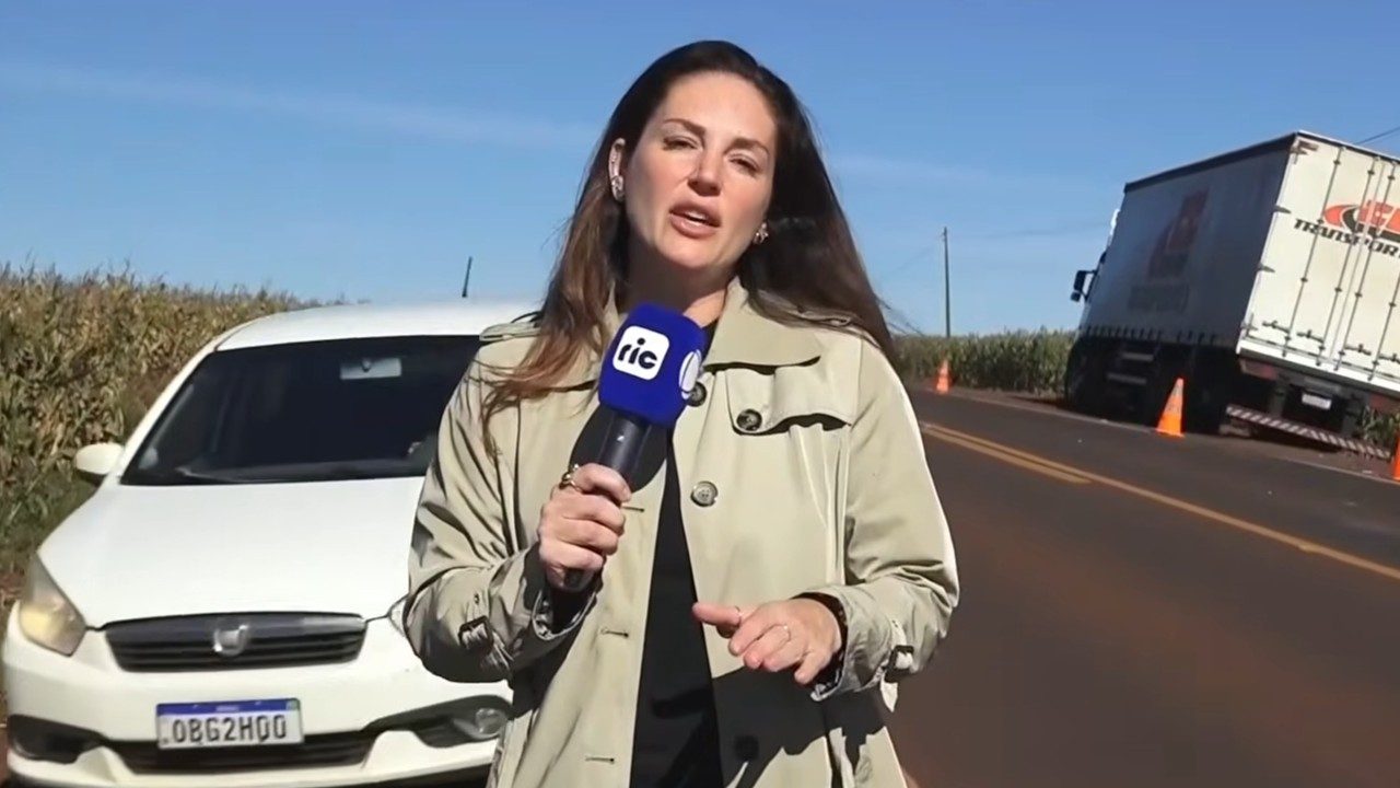 Carolina Barufatti com microfone da Ric TV reporta à beira de rodovia com carro e caminhão ao fundo no Balanço Geral