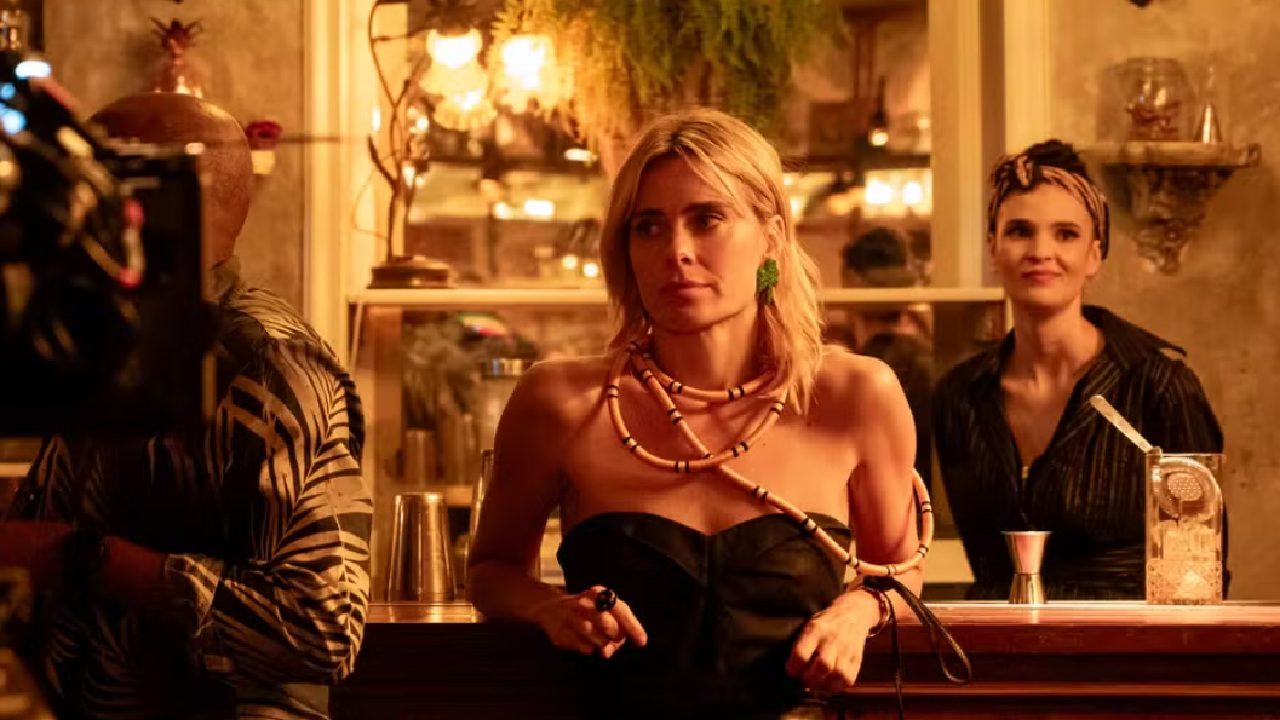 Mulher loira está apoiada no balcão de um bar com iluminação quente e ambiente aconchegante. Ela veste um top preto tomara que caia e acessórios marcantes, como colar longo de contas e brincos verdes. Ao fundo, prateleiras com garrafas, plantas e objetos decorativos compõem o cenário, enquanto outra mulher, atrás do balcão, observa a cena sorrindo.