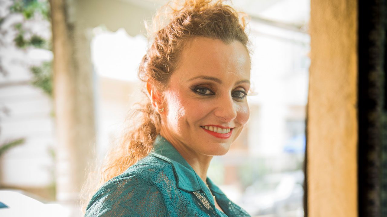 Carolyna Aguiar caracterizada como Lurdinha sorrindo em cenário da novela Totalmente Demais