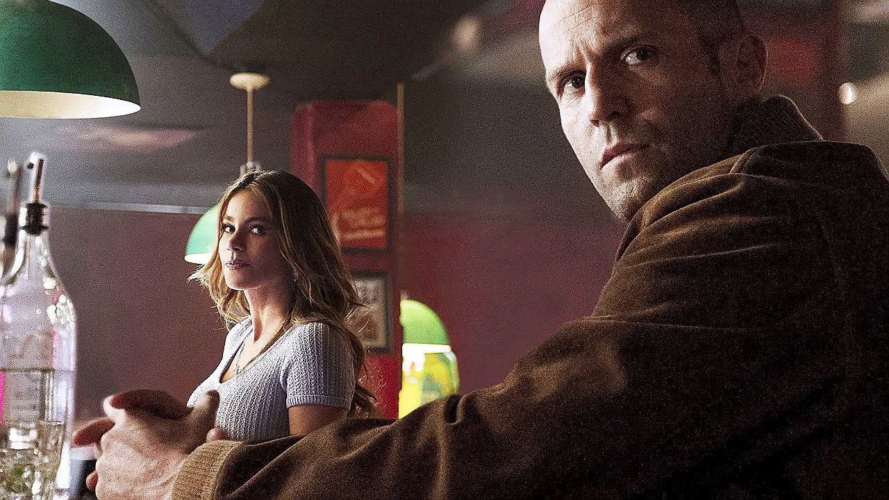 Jason Statham e personagem feminina observam algo fora de cena em bar iluminado em imagem promocional de filme de ação
