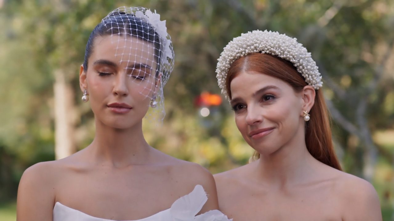 Lorena e Juquinha, duas mulheres com vestidos de noiva e acessórios de pérolas, sorriem durante cerimônia de casamento ao ar livre em cena da novela Três Graças.