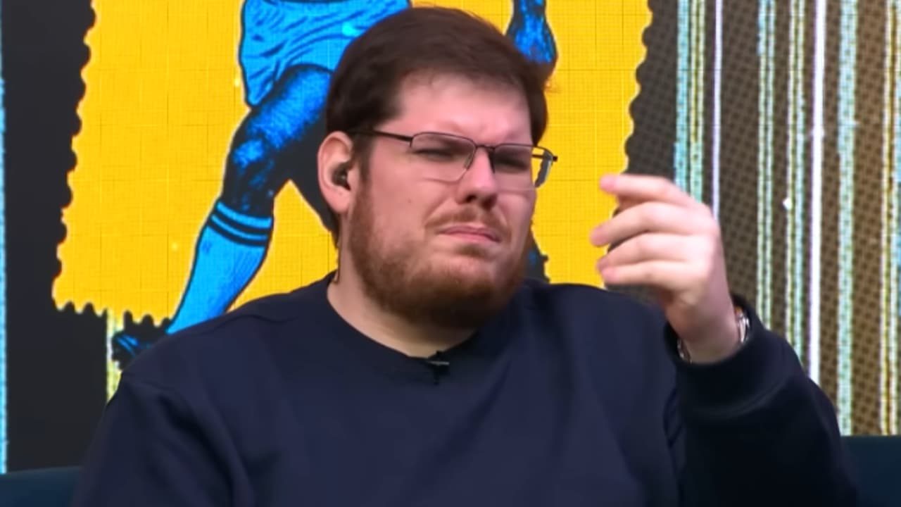 Casimiro Miguel de óculos e moletom escuro faz gesto com a mão durante participação em programa com painel colorido ao fundo
