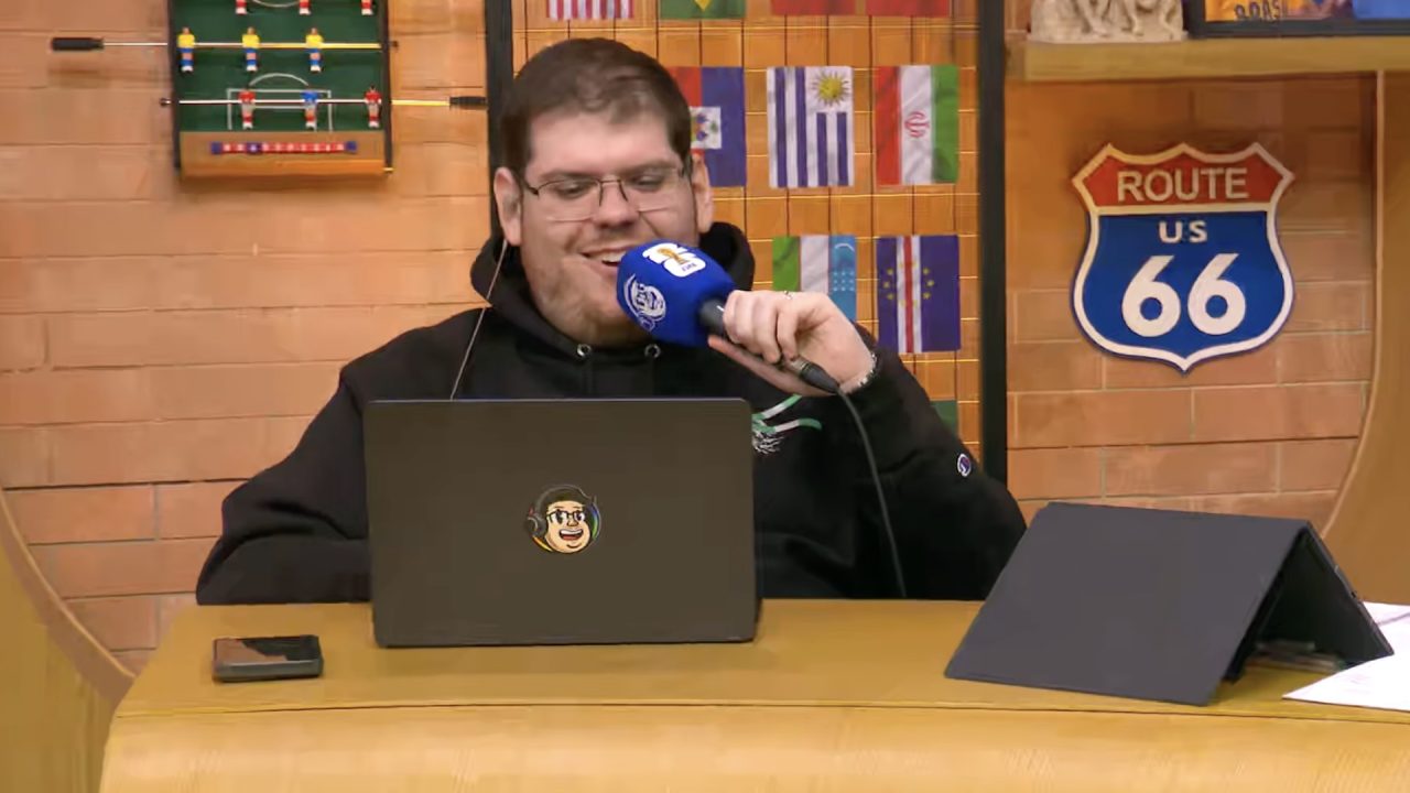 Casimiro Miguel, de moletom preto, sorri com microfone diante de notebook no estúdio da CazéTV