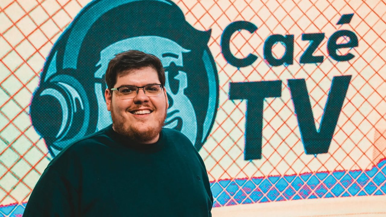 Casimiro Miguel, de óculos e blusa preta, sorri diante de painel com logotipo e nome da CazéTV