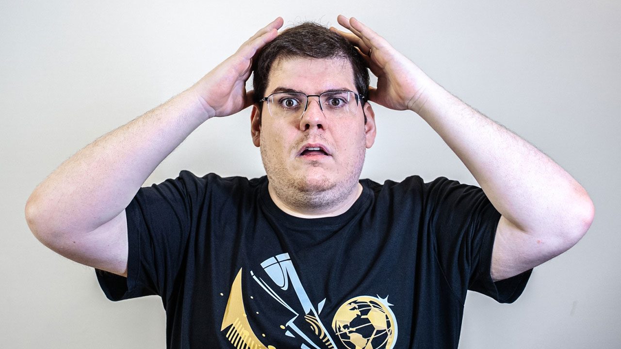 Casimiro Miguel de óculos e camiseta preta com estampa dourada levando as mãos à cabeça em expressão de surpresa