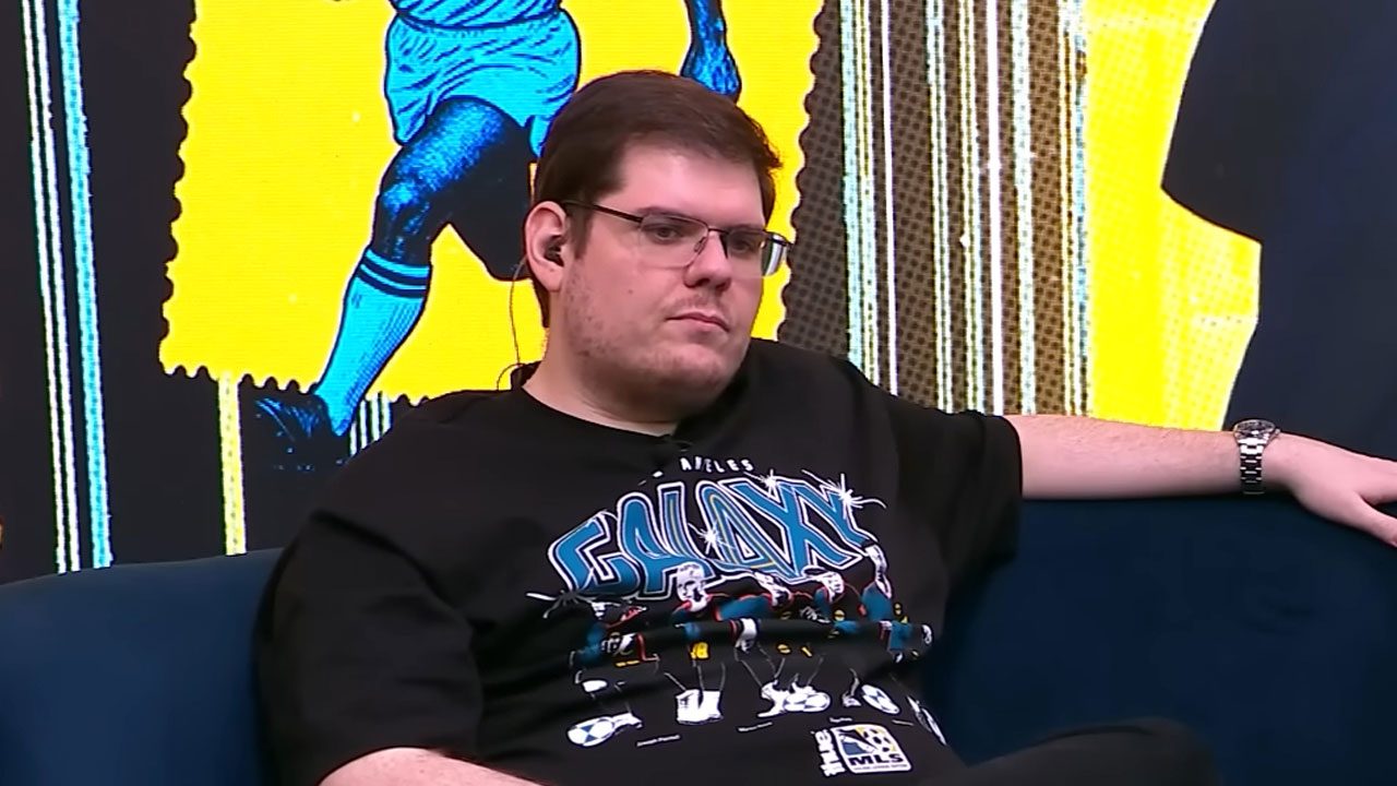 Casimiro Miguel de óculos e camiseta preta com estampa Galaxy sentado em sofá azul durante transmissão ao vivo da Cazé TV