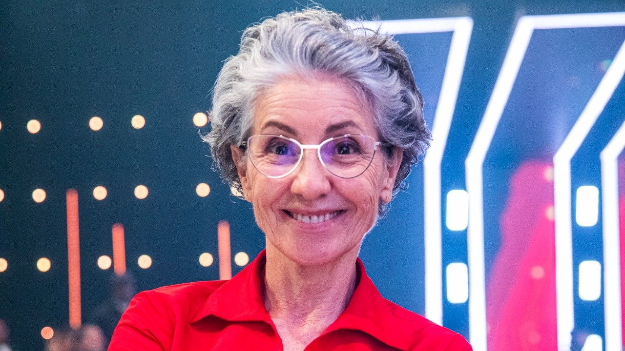 Cássia Kis sorri para a câmera em estúdio de TV com iluminação cênica ao fundo usando camisa vermelha e óculos de armação clara