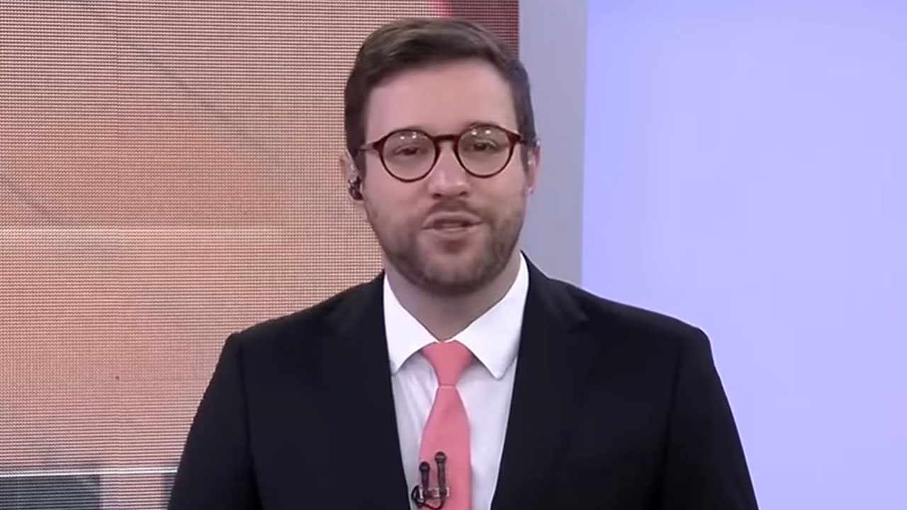 Cassius Zeilmann usa terno preto e gravata rosa enquanto apresenta jornal no estúdio do BandNews TV com óculos de grau redondos