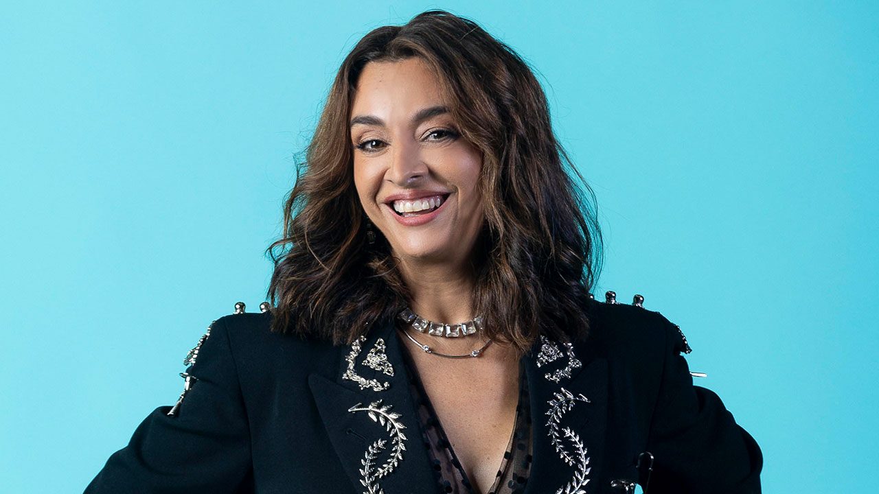 Catia Fonseca sorrindo com blazer preto bordado e colar prateado diante de fundo azul