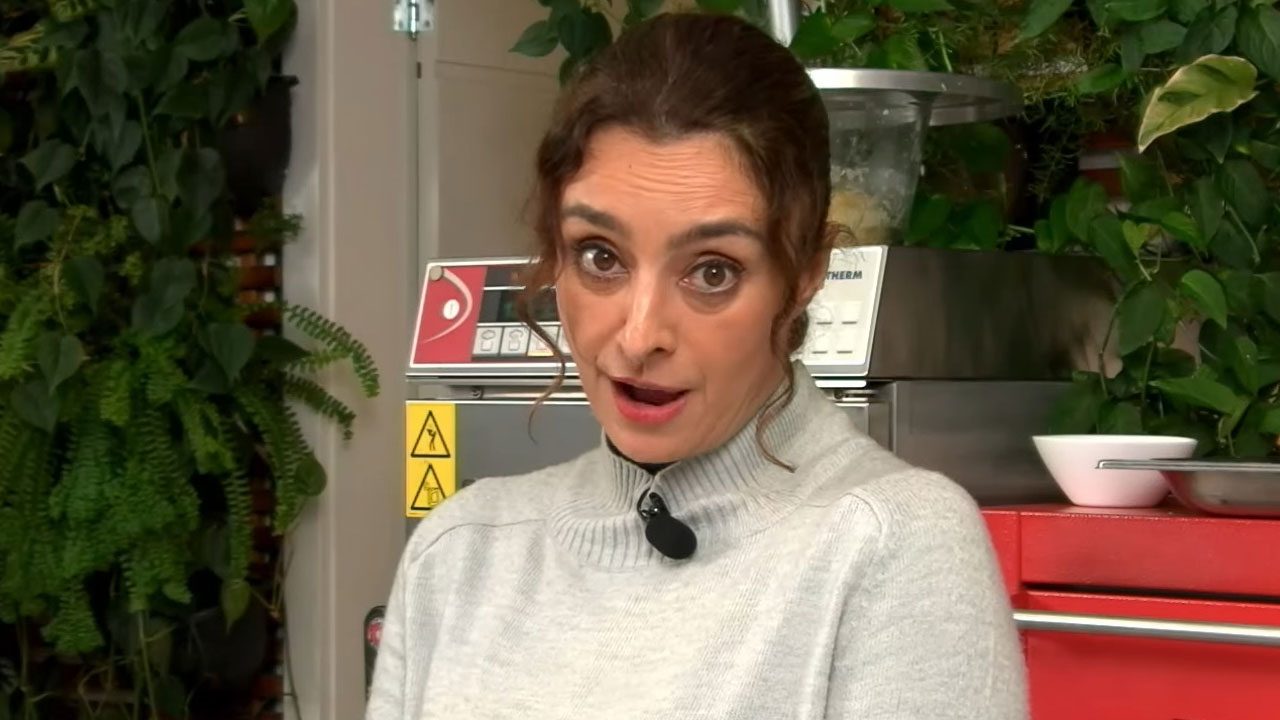 A apresentadora Catia Fonseca com suéter cinza e cabelo preso fala com expressão de surpresa na cozinha de sua casa com plantas ao fundo