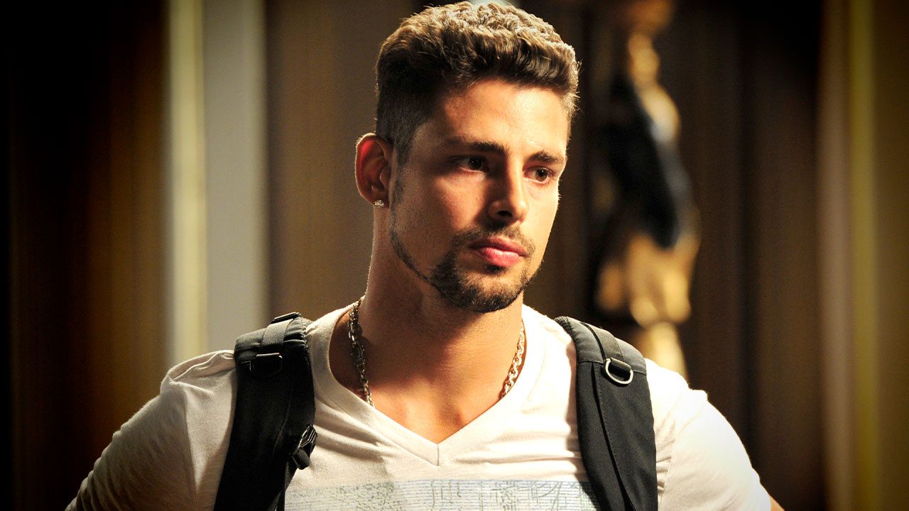 Cauã Reymond caracterizado como Jorginho em Avenida Brasil usando camiseta branca e mochila preta no ombro