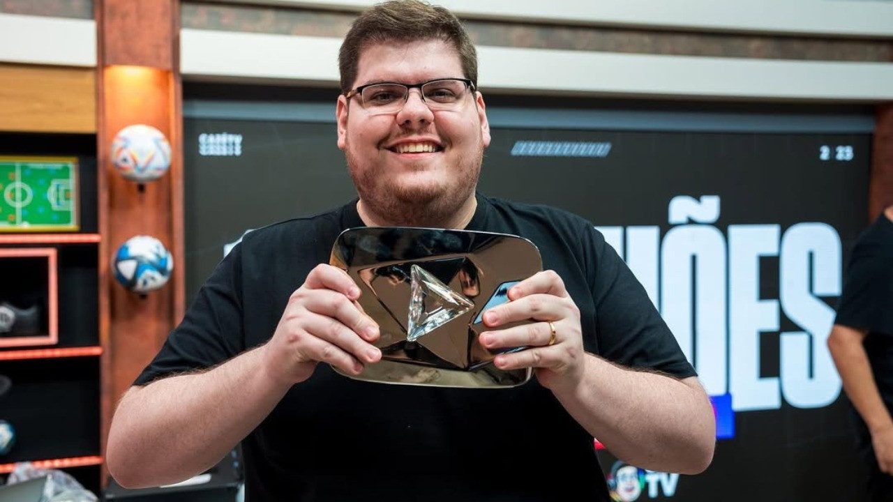 Homem sorridente segura o troféu Silver Play Button do YouTube, usado para premiar criadores de conteúdo, em um estúdio decorado com bolas de futebol e telão ao fundo. A cena transmite clima de comemoração e conquista profissional