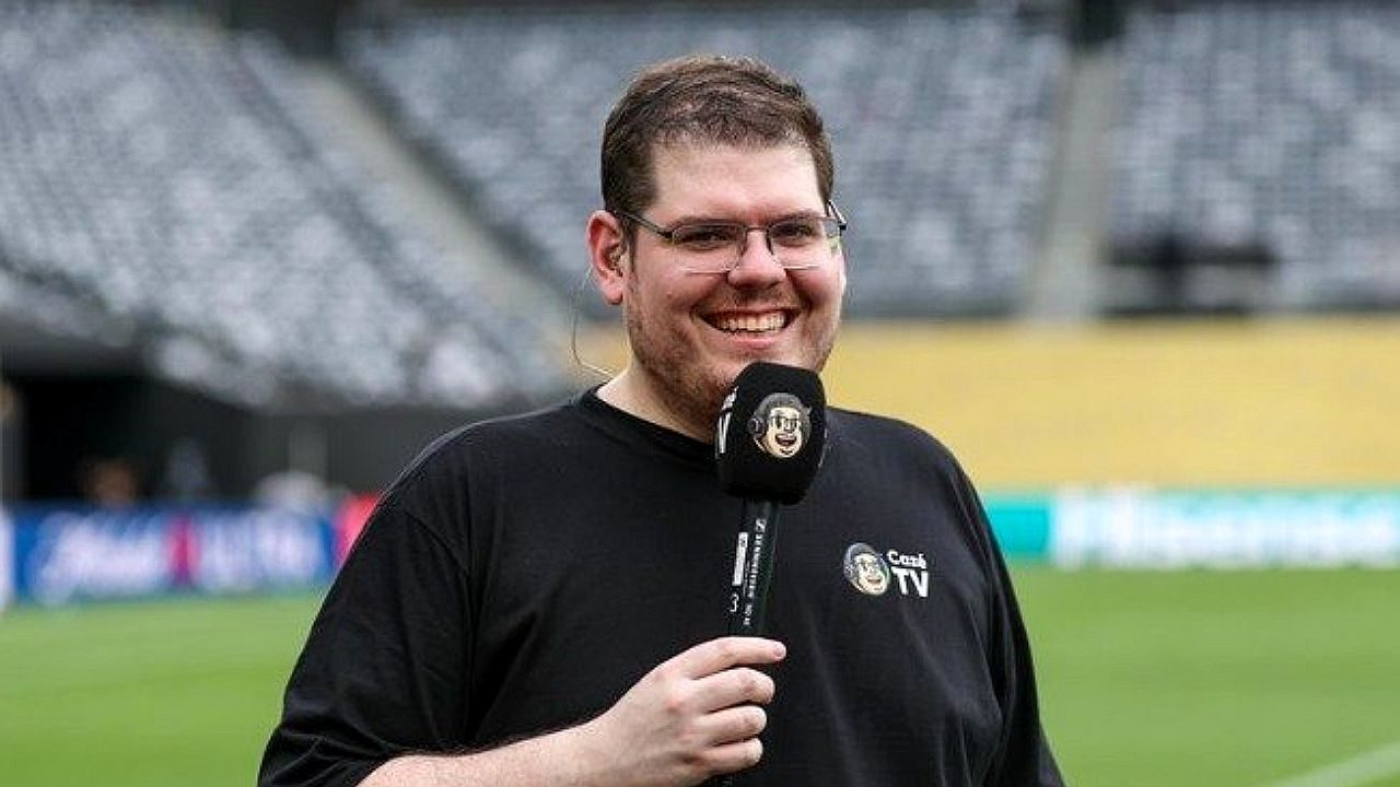 Repórter esportivo sorri no gramado de estádio segurando microfone da Cazé TV