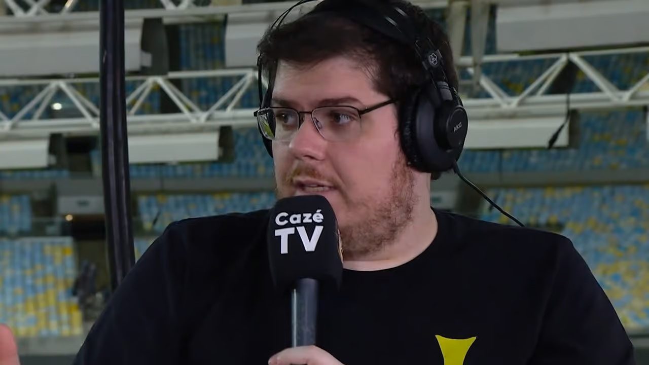 Comentarista sentado em estúdio improvisado dentro de um estádio fala ao microfone enquanto usa fones de ouvido grandes. Ele veste camiseta preta com estampa amarela e segura um microfone identificado como “Cazé TV”, com as arquibancadas desfocadas ao fundo.